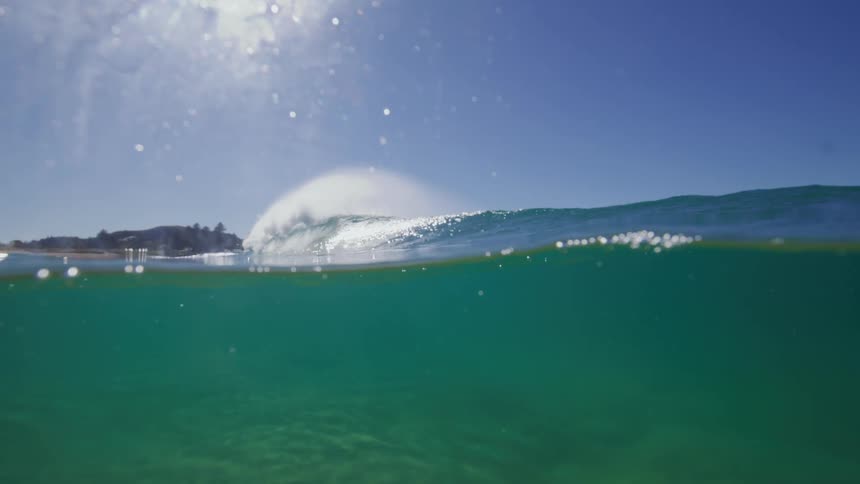A serene over-under shot captures a perfect wave cresting in slow motion before submerging the camera into the clear blue water