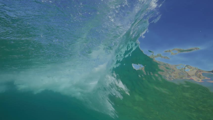 A dramatic underwater slow-motion shot captures a turquoise ocean wave building and breaking over a surfboard