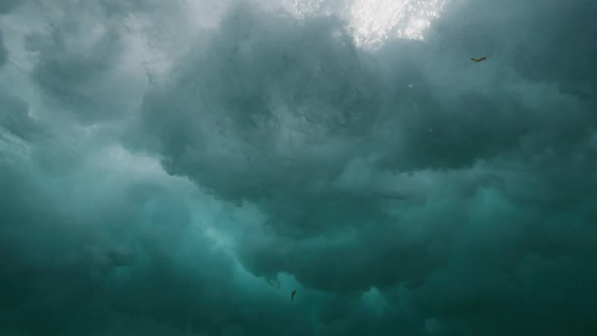 A dramatic underwater slow motion view of a powerful turquoise ocean wave as it curls and breaks, creating a turbulent vortex of bubbles and foam