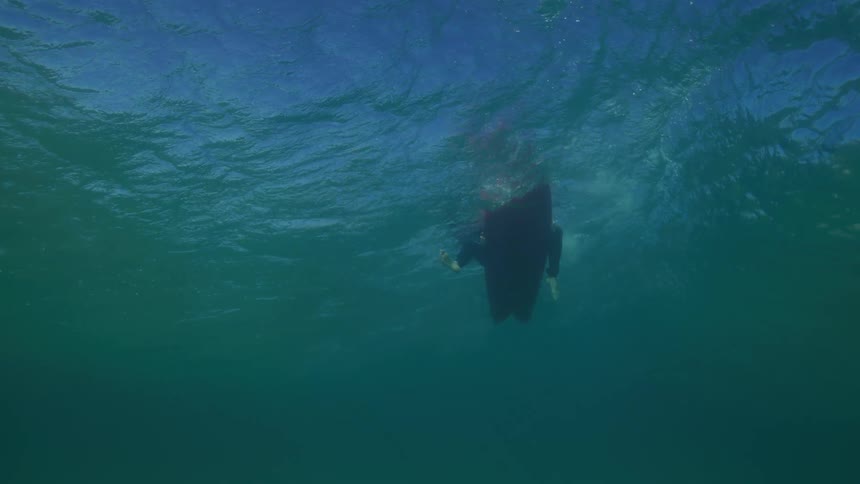 An underwater view shows a surfer in a wetsuit lying on their board, patiently waiting for a wave in the calm ocean