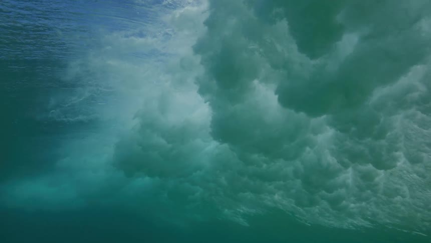 A stunning slow-motion underwater view captures a powerful ocean wave as it forms and crashes, creating a turbulent cloud of whitewater and bubbles