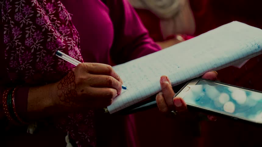 A close-up shot of a woman with a henna tattoo on her hand writing notes in a spiral notebook while holding a smartphone