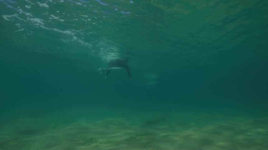 An underwater perspective of a surfer in a wetsuit patiently waiting on their board in the calm, sunlit ocean