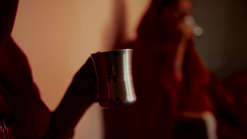 An Indian woman holds a traditional steel glass in her hand while another woman sits in the background