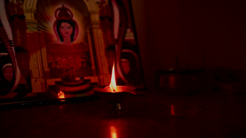 A solitary flame from a Hindu oil lamp flickers in the dark, illuminating a sacred portrait and creating a serene, meditative atmosphere