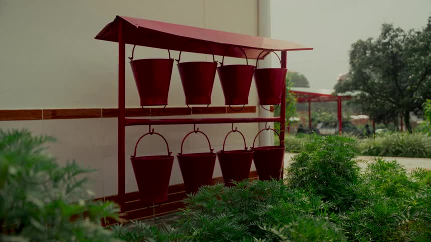 A red fire bucket stand with eight hanging buckets is positioned next to a building wall surrounded by lush green bushes