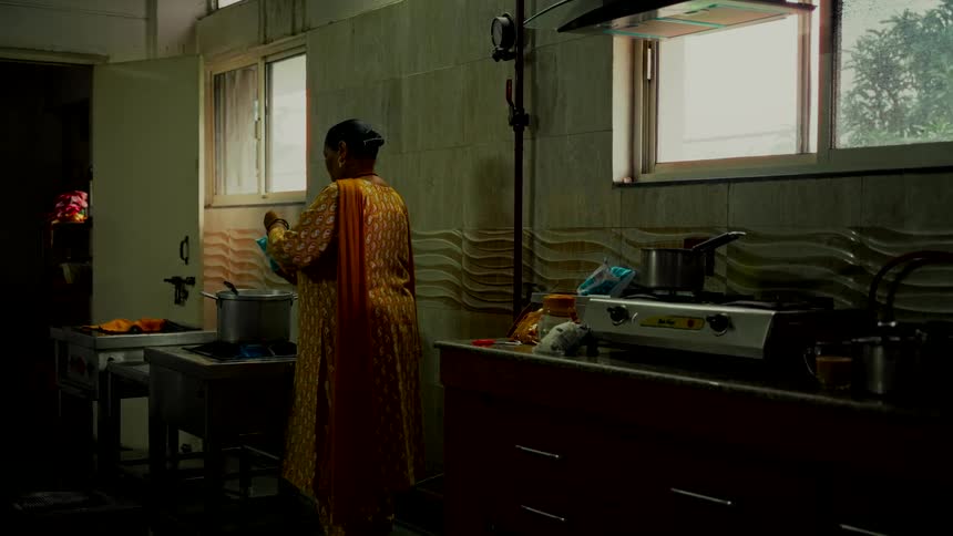 An Indian woman in a yellow salwar kameez prepares a meal in a dimly lit kitchen, pouring milk into a steaming pot on the stove