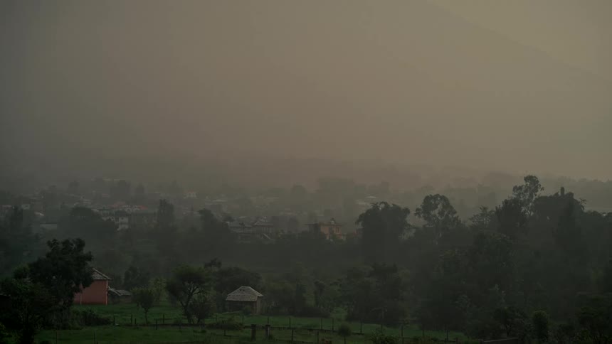 A wide static shot shows a dense, yellowish fog settling over a quiet rural town in a lush, green mountain valley