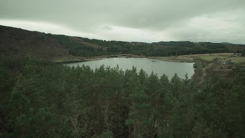 An aerial shot flies over a dense pine forest, revealing a serene loch surrounded by hills on a moody, overcast day