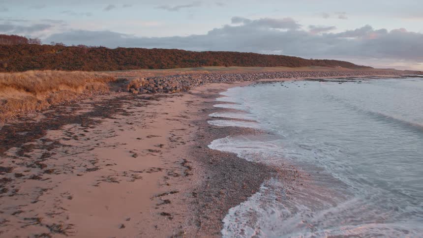Gentle ocean waves in slow motion wash up on a sandy coastline with dunes at sunset