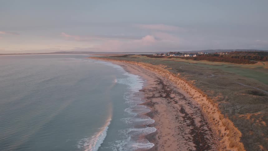 A serene aerial view flying backward along a beautiful coastline at sunset, with calm waves washing onto the sandy beach next to grassy dunes
