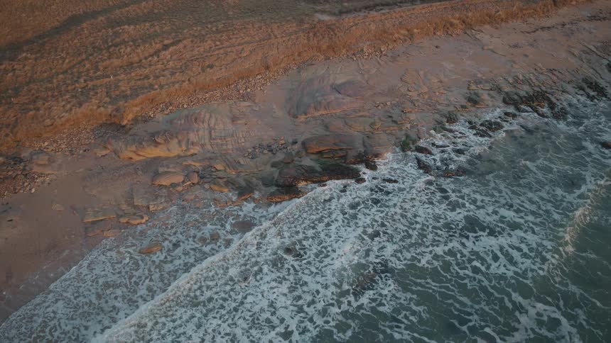 A stunning slow aerial descent shows gentle waves breaking on a rocky shoreline during a beautiful golden hour