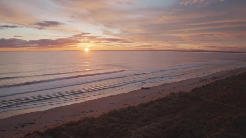 An aerial drone flies over grassy sand dunes toward a calm ocean beach during a beautiful and serene sunset