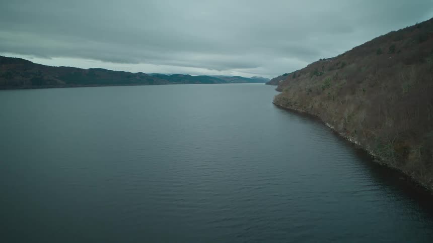 A serene and moody aerial view of a vast loch surrounded by rolling hills under a cloudy winter sky