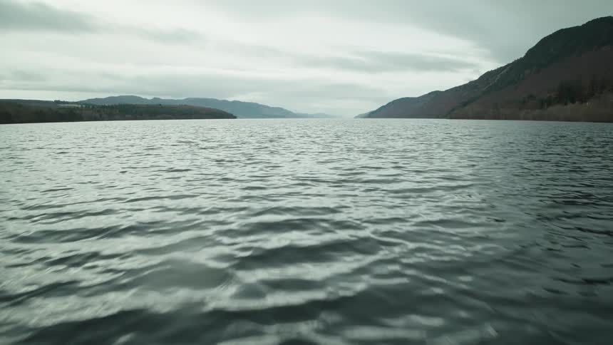 A low, gliding shot reveals the vast, dark waters of a tranquil loch surrounded by hills under a cloudy sky