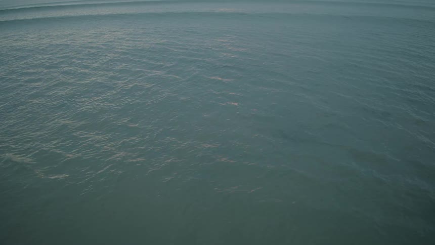 The calm surface of the ocean gently ripples as slow-moving waves roll towards the shore at dusk