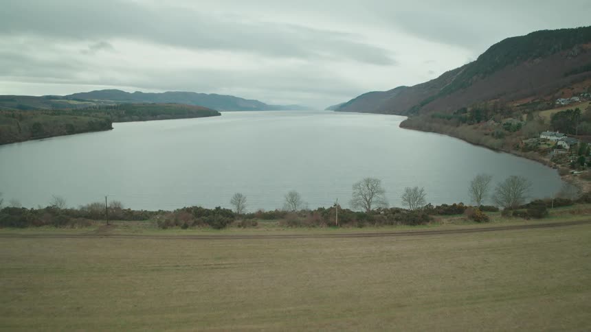 An aerial view rises over pine trees reflecting in the water to reveal the vast, moody expanse of Loch Ness in the Scottish Highlands