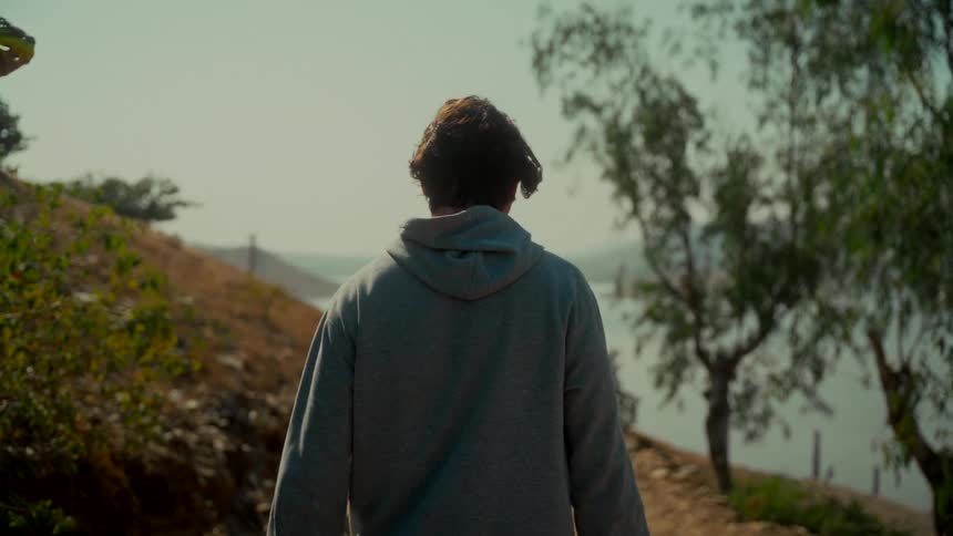 A young man in a grey hoodie walks in slow motion down a dusty path on a hill overlooking a lake