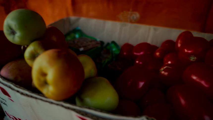 A cinematic close-up reveals a cardboard box filled with vibrant red tomatoes, fresh apples, and oranges at a local market