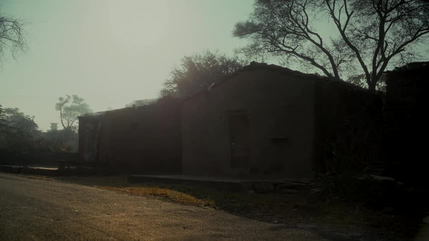 Smoke rises from a rustic mud house on a hazy morning in a quiet rural village