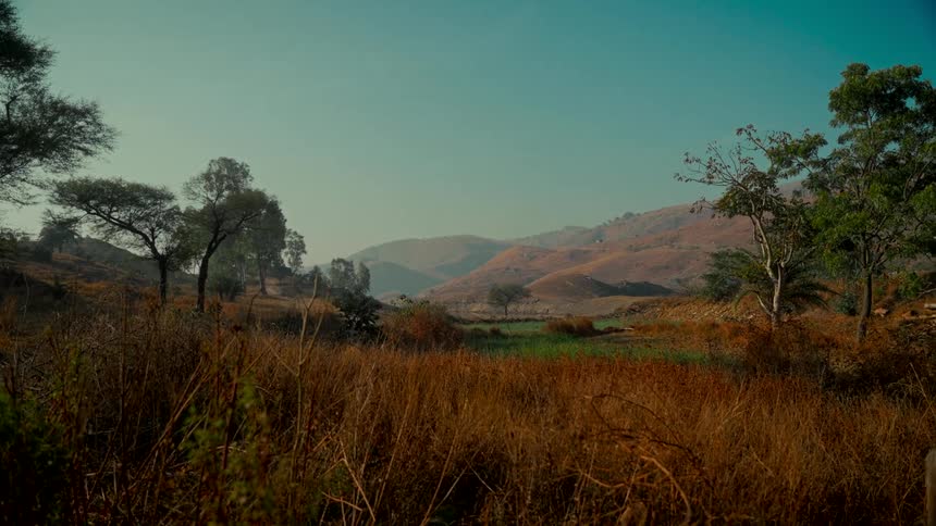 A serene, static landscape of a rural valley with dry grass in the foreground and hazy, rolling hills in the distance under a clear sky
