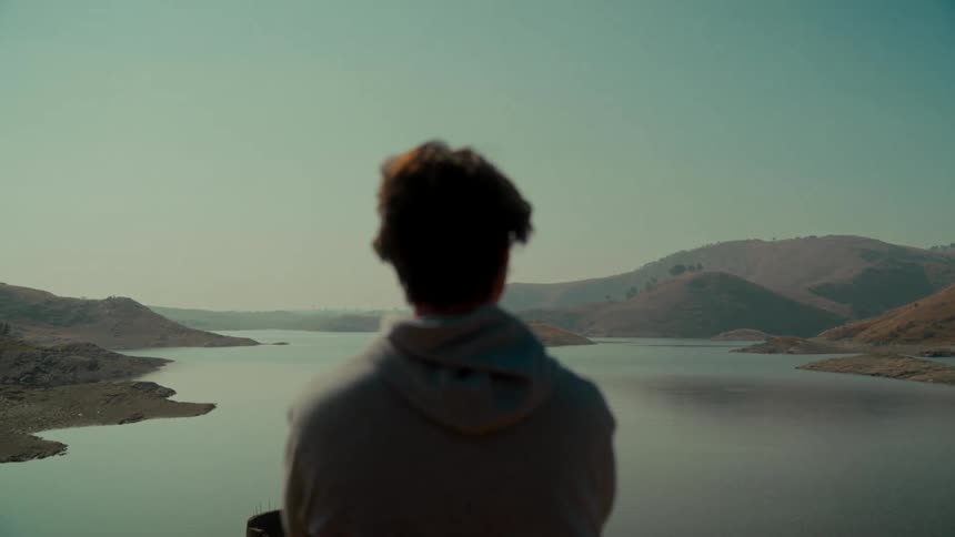 An over-the-shoulder shot shows a pensive person in a hoodie looking out at a serene lake surrounded by dry, rolling hills