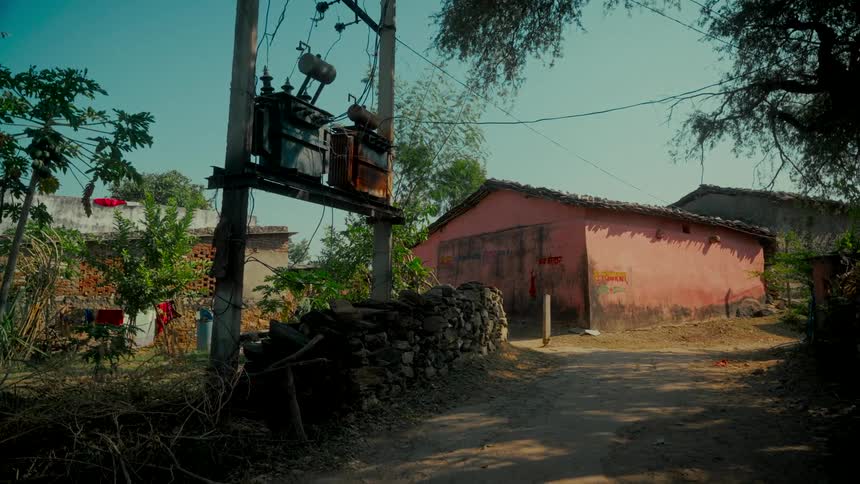An old electrical transformer sits on a utility pole next to a weathered pink building in a quiet rural village
