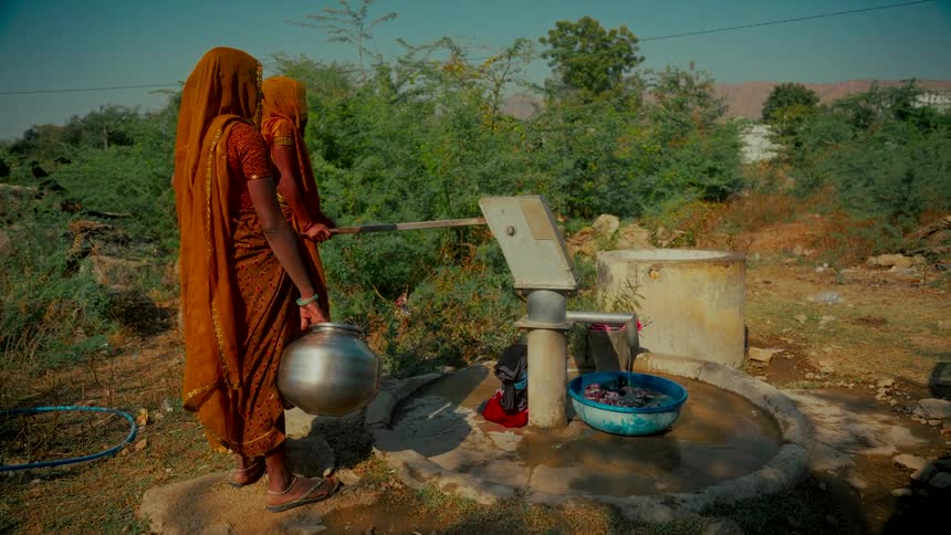 Two Indian women in traditional orange sarees collect fresh water from a hand pump in a rural village