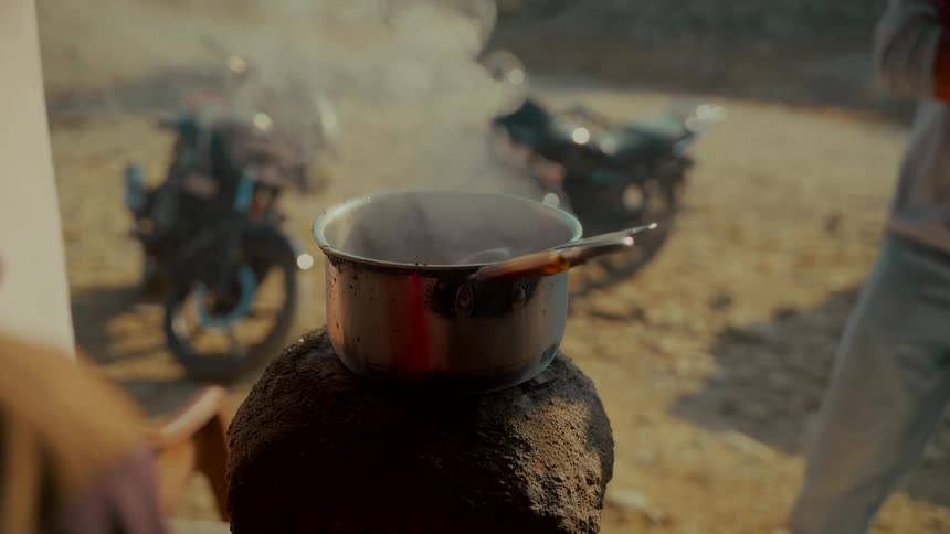 Over-the-shoulder slow-motion shot of a person stirring steaming chai tea in a pot on a rustic stove
