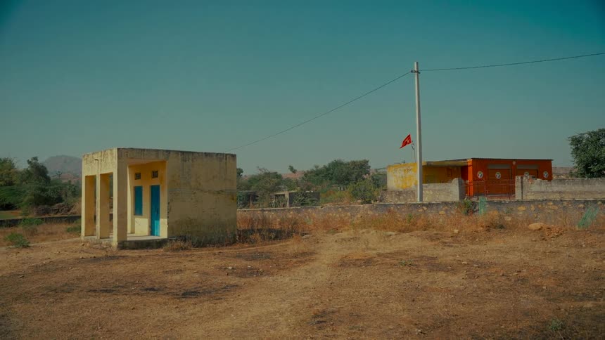 A static wide shot shows a small red flag fluttering in the wind in a quiet, arid rural village