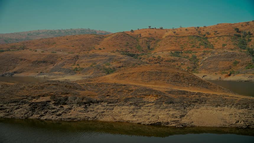 A tranquil and arid landscape of rolling brown hills surrounding a calm lake under a clear sky
