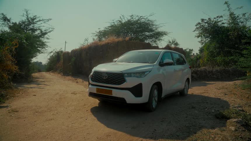 A white SUV drives slowly down a dusty, rural road in the countryside on a sunny day