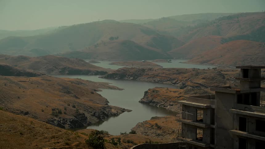 A vast and hazy mountain landscape with a river winding through the arid hills next to an unfinished building
