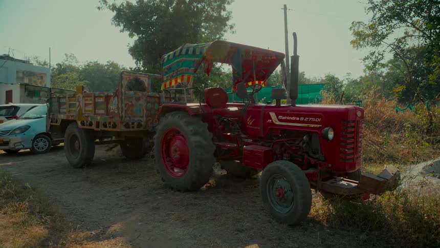 An old red tractor with a decorated trailer is parked on a dirt road in a rural village setting