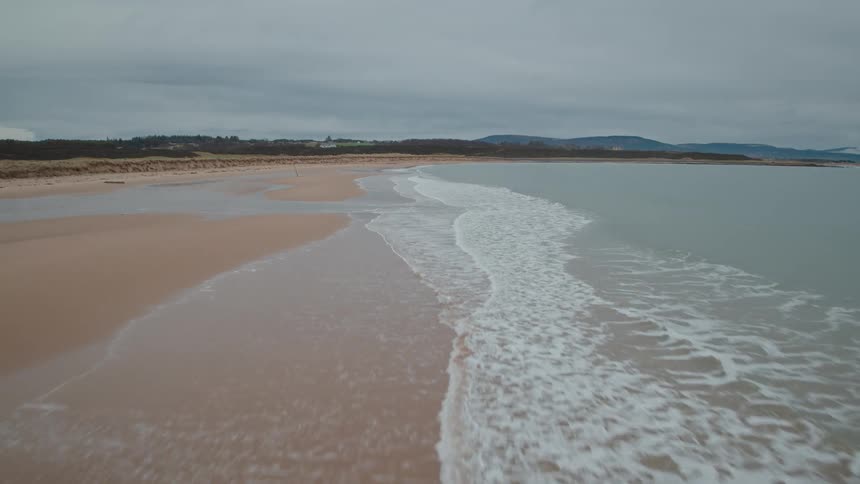A serene aerial view pulls back over waves gently lapping against a deserted sandy beach on a tranquil, overcast day