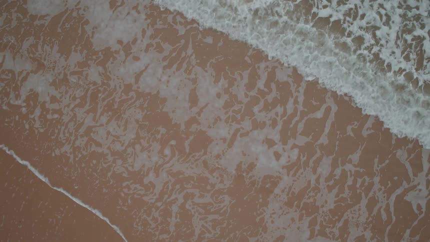 A mesmerizing top-down aerial view captures the gentle rhythm of foamy ocean waves washing over a sandy shore in slow motion