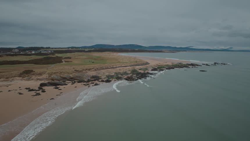 An aerial view rises over a vast, sandy beach on a cloudy day, revealing the full extent of the dramatic coastline
