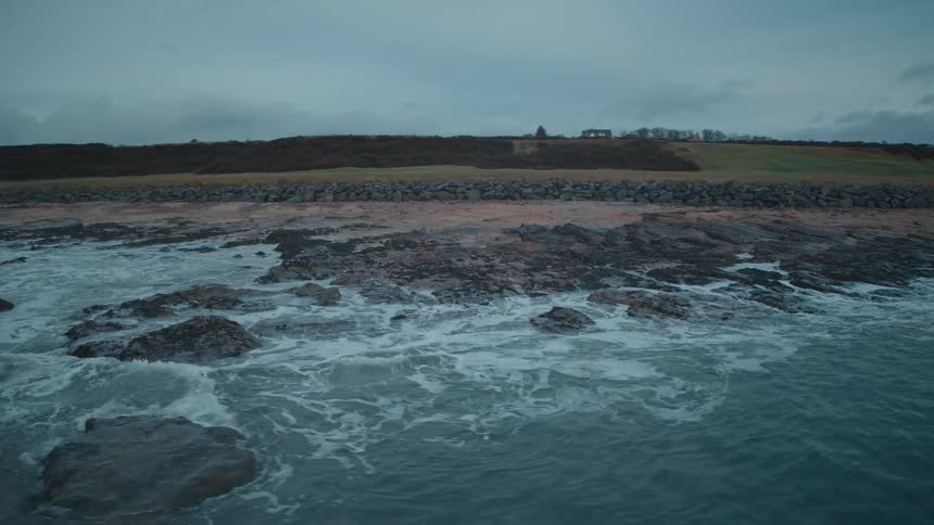 A slow-motion aerial shot captures the ocean waves gently washing over a rocky and sandy beach during a moody twilight