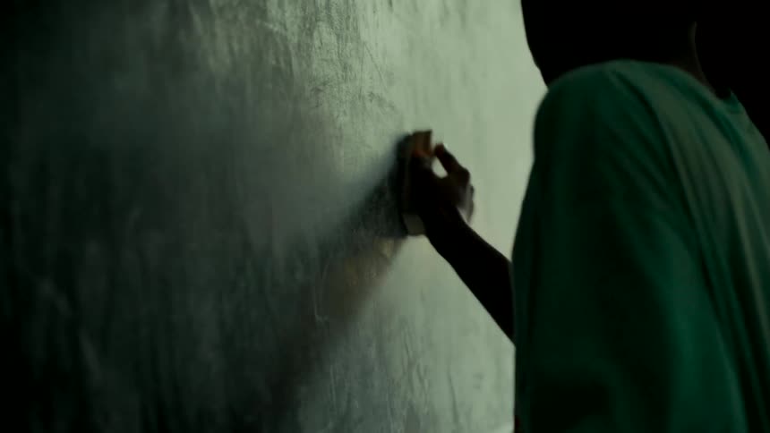 A silhouetted student erases a dusty chalkboard in a dimly lit classroom