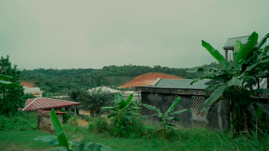A quiet, rural village sits nestled amongst lush green hills and tropical plants on a cloudy, overcast day