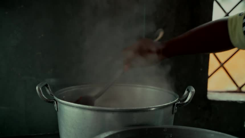 A person stirs a large aluminum pot of hot, steaming food in a rustic kitchen with a ladle
