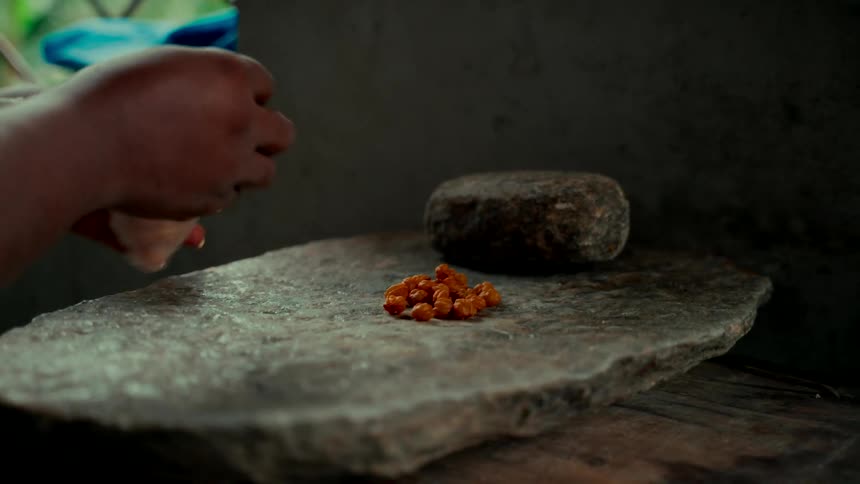 A person's hands slowly pour small orange berries onto a traditional stone grinding slab in a rustic kitchen