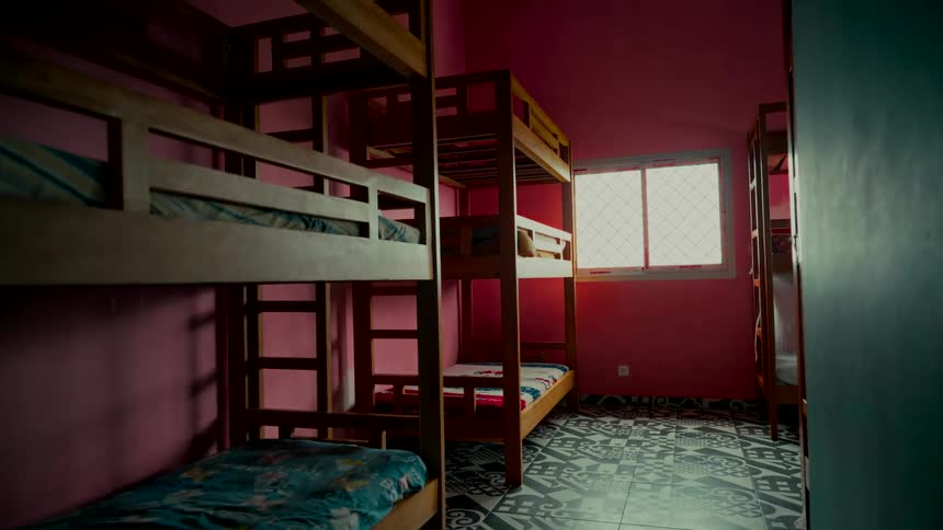 A static wide shot of a dimly lit, empty hostel room with wooden bunk beds, pink walls, and decorative tile flooring