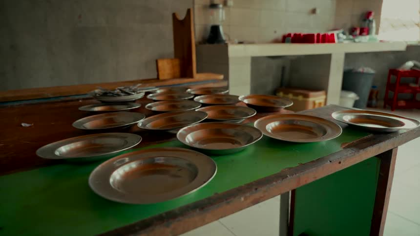 Empty metal plates are neatly arranged on a rustic table in a simple institutional kitchen