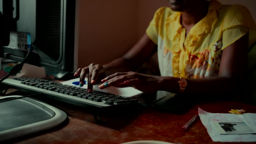 Close-up shot of a Black woman's hands with colorful painted nails typing on a computer keyboard at a wooden desk