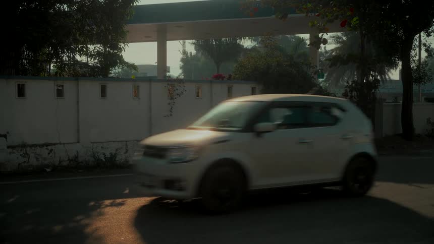 A white car drives slowly down a sunlit road with a gas station in the background