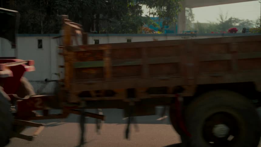 Slow motion side view of a red tractor pulling a large rusty trailer on a paved road during the day