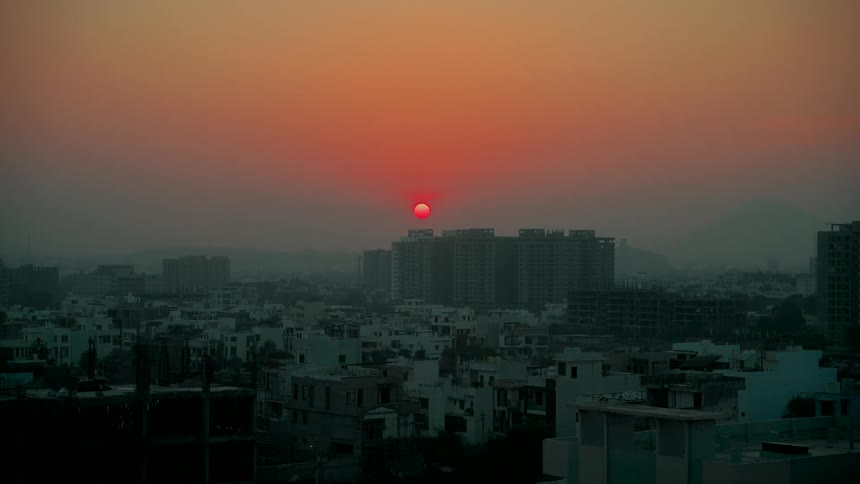 The red sun glows in the hazy sky during a warm sunset over a sprawling urban cityscape