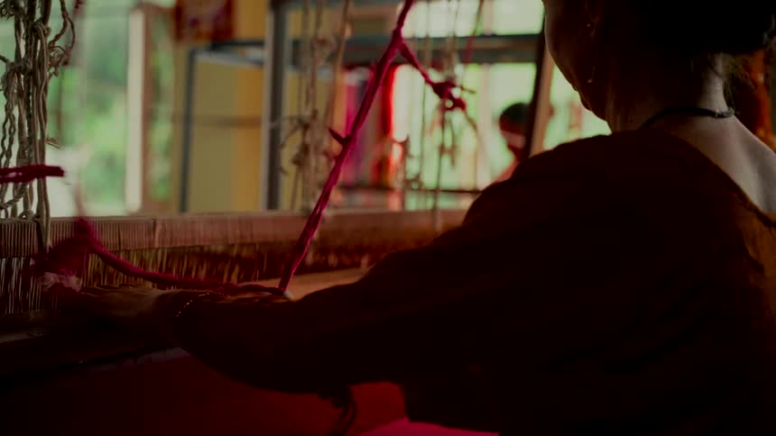 A silhouetted woman skillfully weaves colorful fabric on a traditional wooden handloom inside a rustic workshop