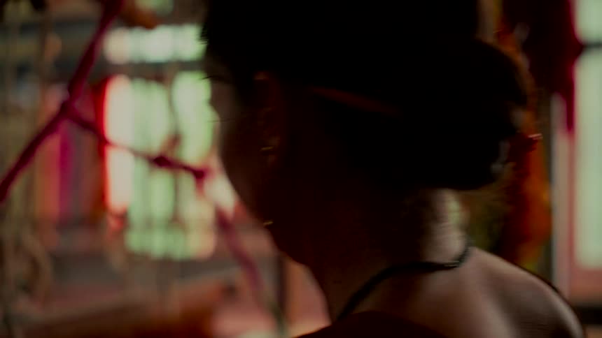 Over-the-shoulder view of a silhouetted woman with her hair in a bun weaving pink threads on a traditional loom